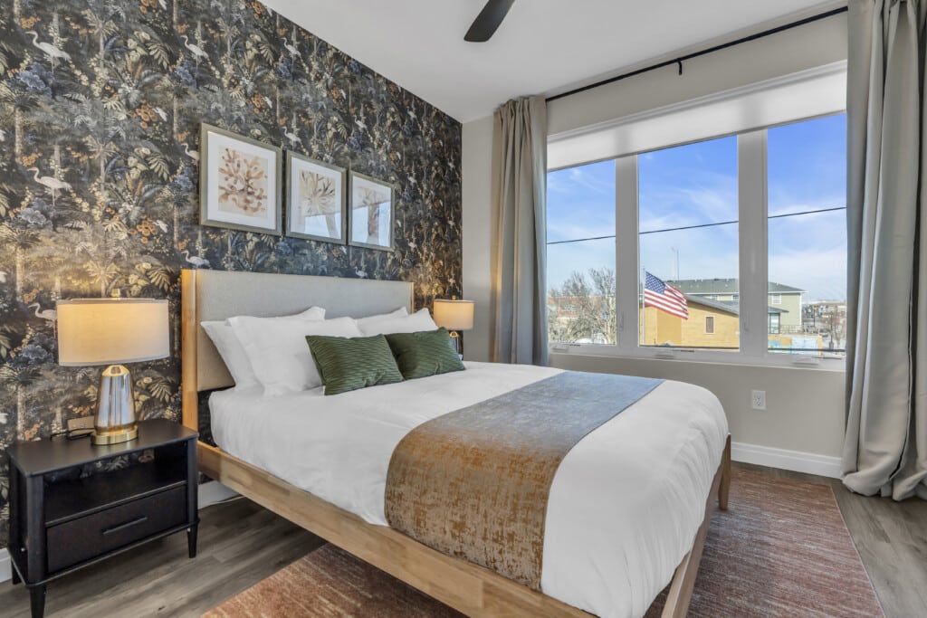 Cozy bedroom with a bed featuring green accent pillows, decorative wall art, and large windows showcasing an American flag outside, reflecting the appeal of Jersey Shore vacation rentals.