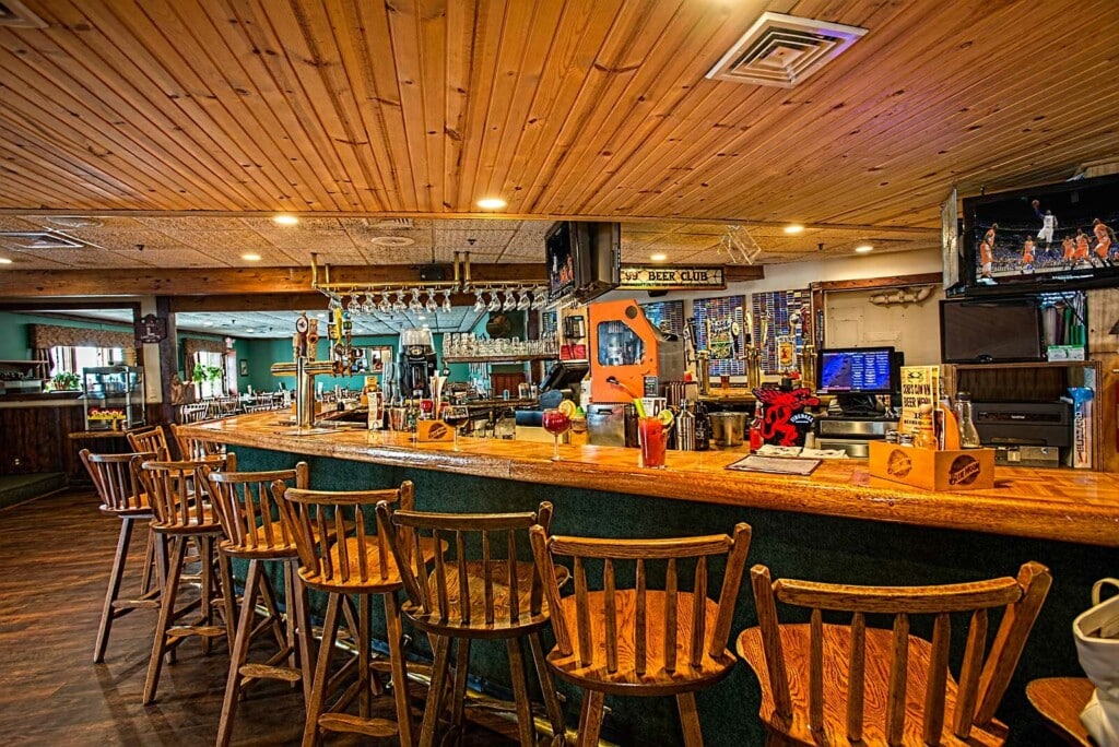 Cozy bar interior with wooden stools and a long counter, featuring a variety of drinks and a welcoming coastal atmosphere, ideal for enjoying casual dining near the beach at Bum Rogers Crabhouse.