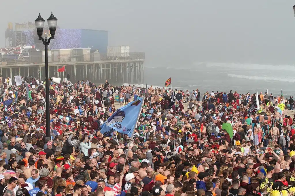 Crowd of participants at the Seaside Heights Polar Bear Plunge event, with people gathered on the beach, some in swimwear, flags waving, and the boardwalk in the background, celebrating and preparing for the plunge.