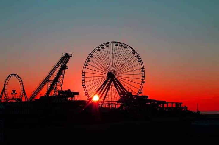 Silhouette of a Ferris wheel and amusement rides against a vibrant sunset, representing the iconic attractions of the Jersey Shore boardwalk experience.
