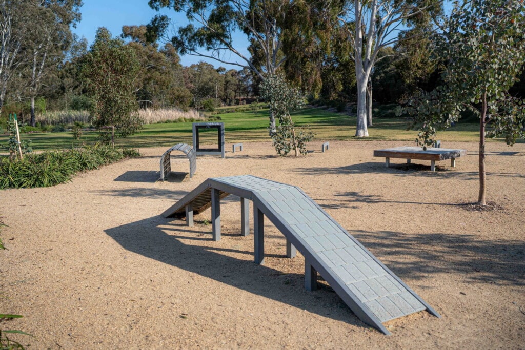 Dog agility equipment in a sandy area of a dog park, featuring a ramp, tunnel, and benches surrounded by trees, ideal for off-leash fun at Fisherman’s Cove Conservation Area.