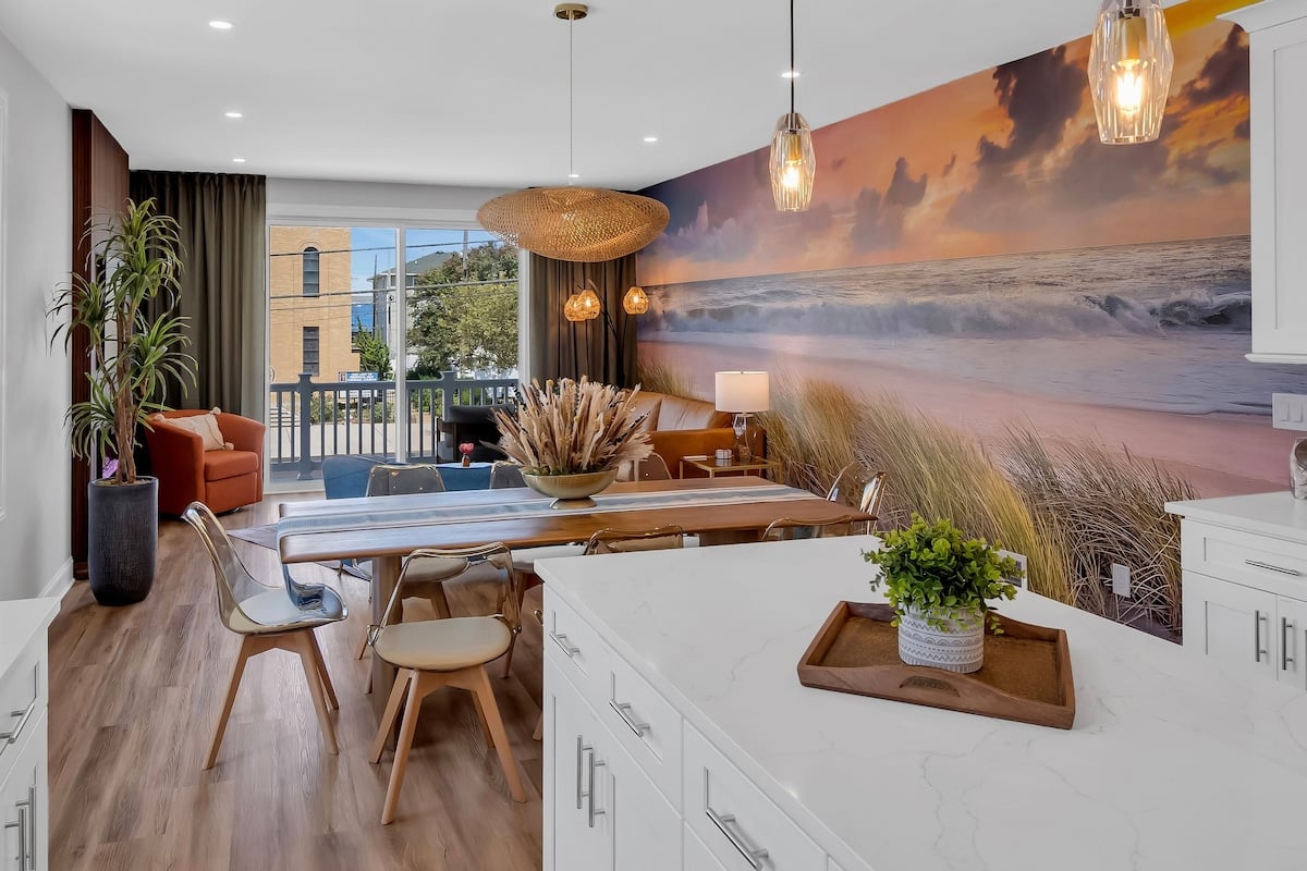 Modern beach-inspired living room featuring a dining area with a wooden table, transparent chairs, and coastal decor, complemented by a mural of a serene ocean landscape at sunset, reflecting the Jersey Shore vacation rental ambiance.