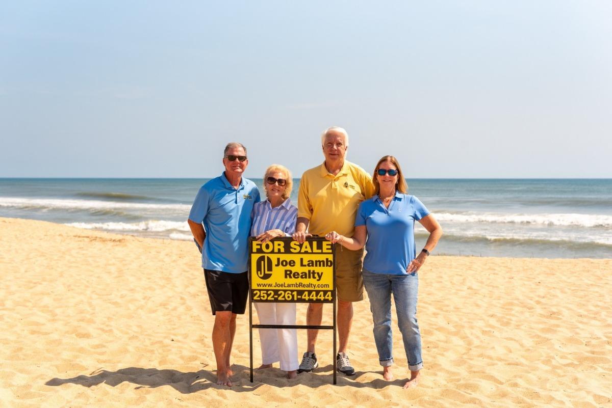4 realtors standing on the beach behind a for sale sign