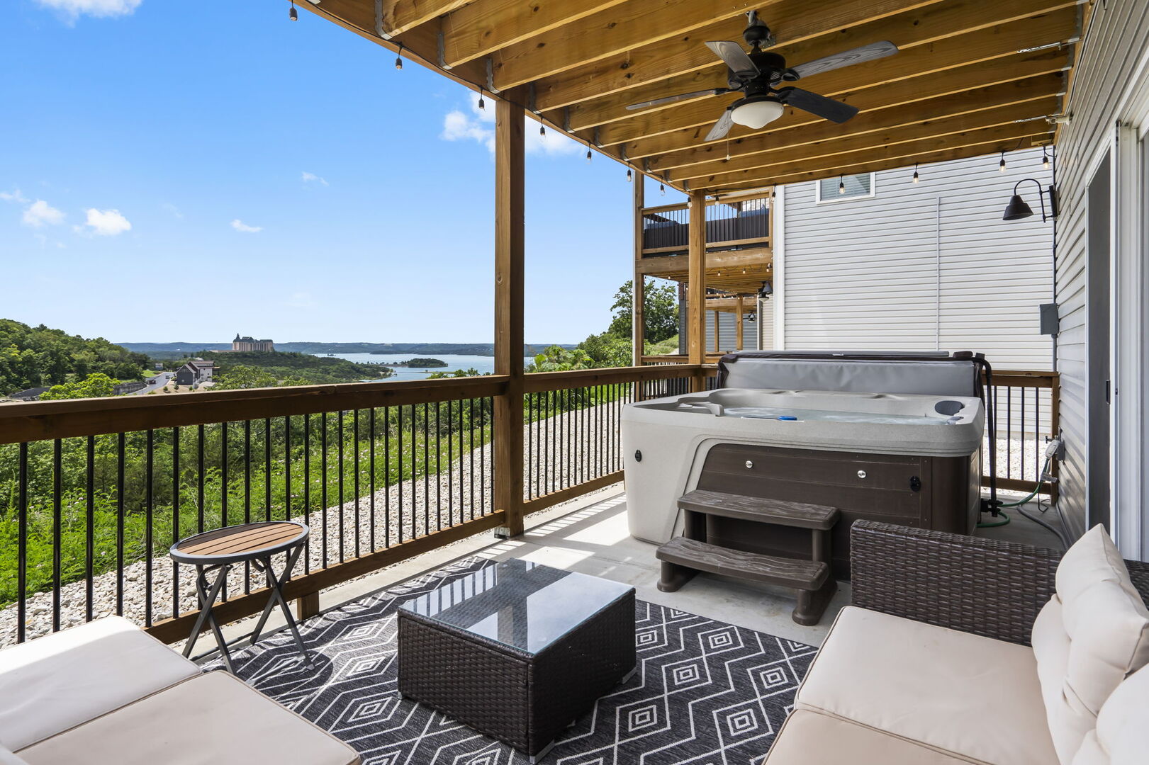 Covered deck with a private hot tub, outdoor seating and a ceiling fan, looking over wooded hills to the lake