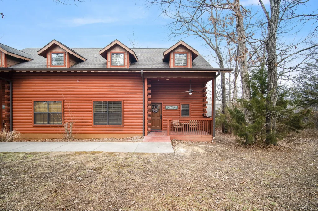 Red log-style cabin with three dormer windows and a covered front porch, set among bare trees