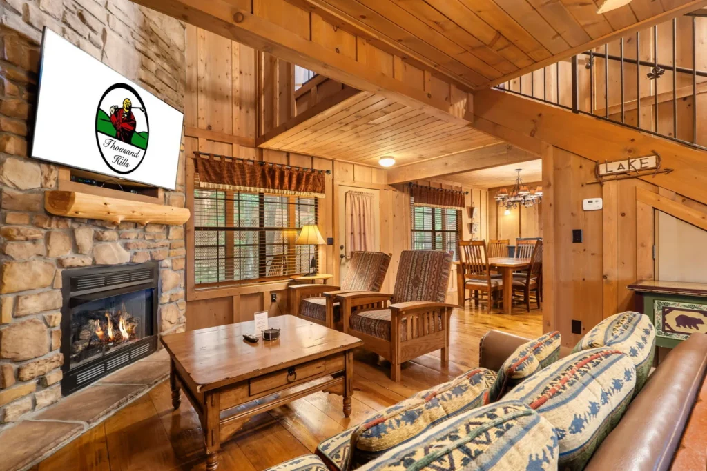 Living room of a Branson cabin rental with a lit gas fireplace set in stone, knotty pine walls and a dining area beyond