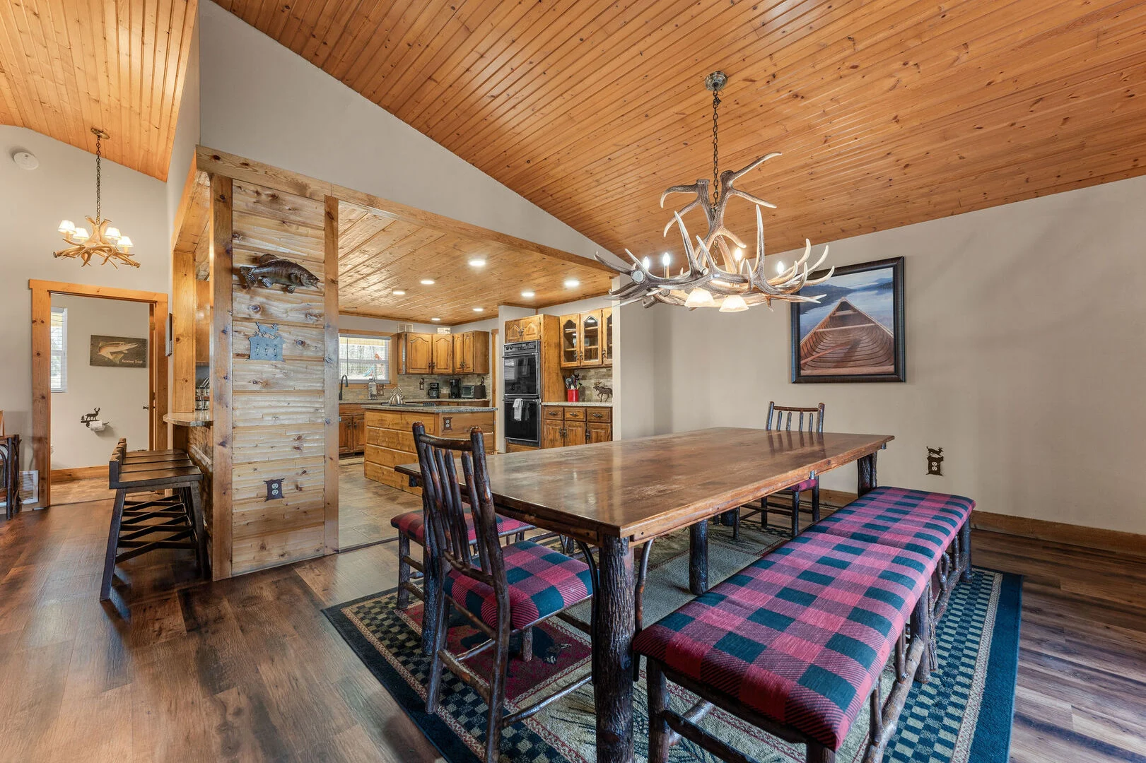 Long farmhouse dining table with plaid benches and an antler chandelier beside the open kitchen of a Branson lodge rental, set up for a Thanksgiving dinner for a big group