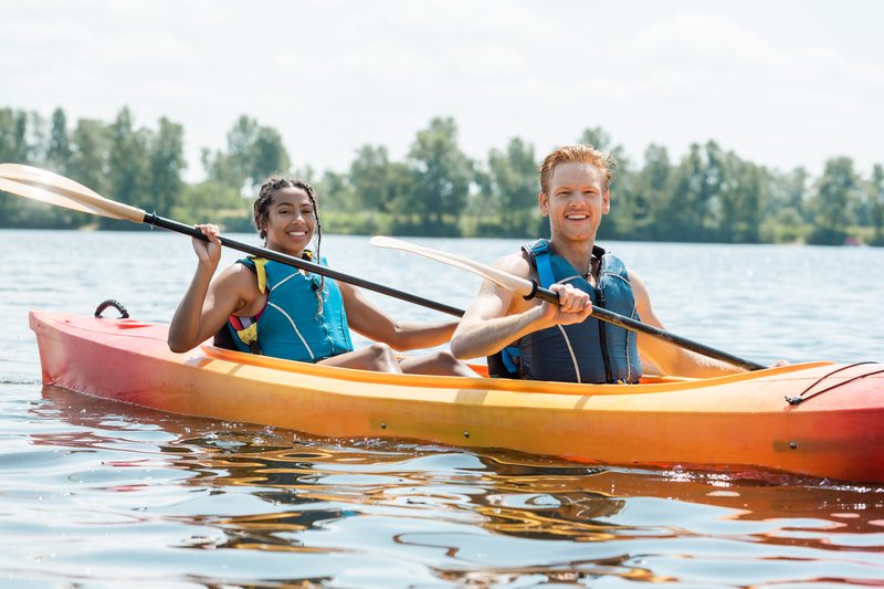 Two paddlers in life vests sharing a red and orange tandem kayak on calm open water under a bright summer sky