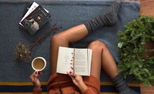 A woman enjoys a book while sipping coffee, immersed in her reading in a cozy setting, with wreath beside her.