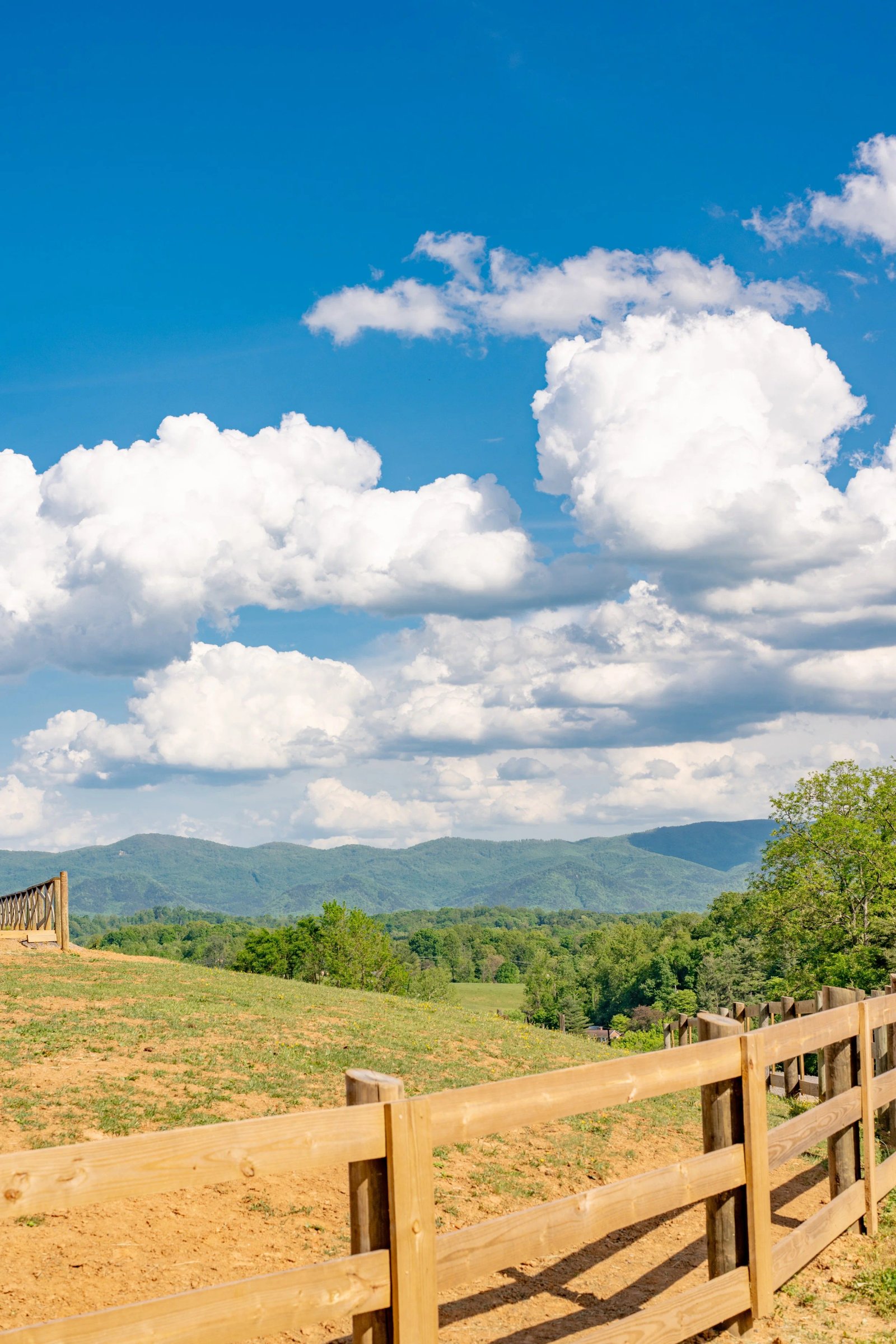 Rural pasture with wooden fencing, green foothills, and dramatic white clouds under a bright blue sky